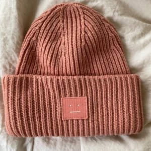 Rosa stickad mössa från Acne Studios - Snygg rosa stickad mössa från Acne Studios med ribbad design och ett broderat ansikte på framsidan. Perfekt för att hålla värmen under kyliga dagar. En stilren accessoar som passar till många outfits.