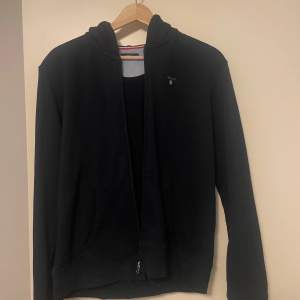 Säljer en stilren svart hoodie från Gant. Den har en dragkedja framtill och en klassisk design med långa ärmar. Perfekt för en avslappnad look.
