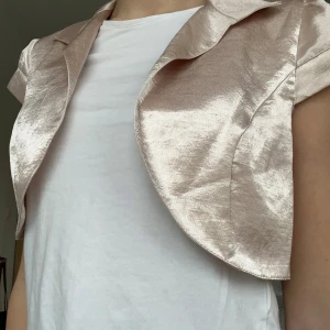 Glansig bolerotopp i beige  - Snygg och glansig bolerotopp i beige med korta ärmar och en elegant krage. Perfekt för att lägga till en touch av glamour till din outfit. 