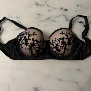 Svart spets-bh med blommönster - Elegant svart bh med vacker spets och blommönster. Den har justerbara axelband och en klassisk hakknäppning bak. Perfekt för en sofistikerad look.