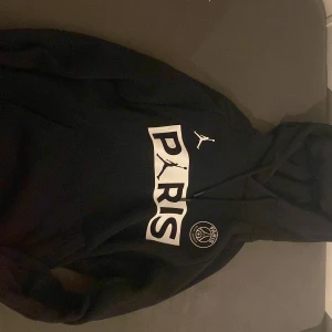 Svart hoodie från Jordan x Paris Saint-Germain - Säljer en svart hoodie från Jordan i samarbete med Paris Saint-Germain. Hoodien har en stor logga med texten 'PARIS' och den ikoniska Jumpman-symbolen. Perfekt för sportiga tillfällen eller som en stilren vardagslook.