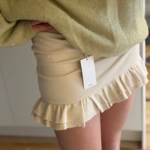Beige kjol med volangdetalj från Mango -  Beige (/off white), kort kjol med figursydd passform och volangdetalj längst ner. Perfekt för både vardag och fest. Gjord av tjockare material, likt en jeanskjol (100% bomull). Enkel att matcha med stickade tröjor eller blusar för en chic look. Storlek:S