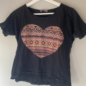 Svart t-shirt med hjärtmönster - Säljer en svart t-shirt med ett stort hjärtmönster i olika färger på framsidan. T-shirten har korta ärmar och en rund halsringning. Perfekt för en avslappnad stil. 🖤