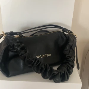 Svart axelväska från Valentino - Snygg svart axelväska från Valentino med rynkad axelrem och dragkedja. Väskan har en elegant design med guldfärgade detaljer och logotyp framtill. Perfekt för att ge en stilren touch till din outfit.