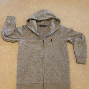 Grå hoodie från Polo Ralph Lauren - Säljer en stilren grå hoodie från Polo Ralph Lauren med dragkedja och klassisk logga på bröstet. Tröjan har långa ärmar och en praktisk huva. Perfekt för en avslappnad stil. Hör av dig vid intresse!
