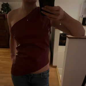Vinröd oneshoulder topp - Snygg vinröd oneshoulder topp i fakeskinn material. Perfekt för en kväll ute. Toppen har en tight passform som framhäver figuren. Aldrig använd!