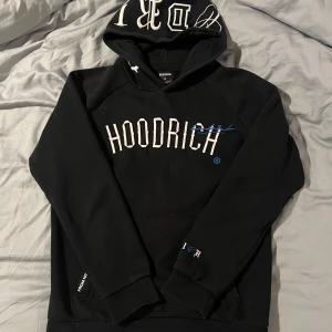 Svart Hoodrich Hoodie - Säljer en svart hoodie från Hoodrich med vit text på framsidan och detaljerad text på baksidan. Hoodien har en stor ficka framtill och en huva med tryck.
