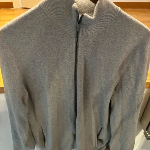 Grå kofta med dragkedja från Massimo Dutti - Snygg och stilren grå kofta från Massimo Dutti med dragkedja framtill. Perfekt för lager-på-lager under kyligare dagar. Den har en hög krage och långa ärmar.