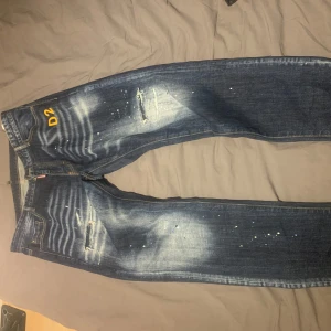 Blå jeans från Dsquared2 - Snygga blå jeans från Dsquared2 med slitna detaljer och coola färgstänk. De har en klassisk femficksdesign och en broderad logga baktill. Perfekta för en trendig look!