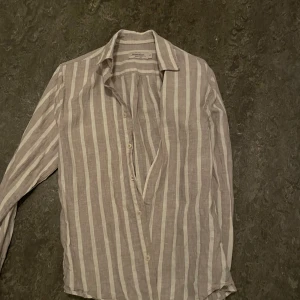 Randig skjorta från Dressmann - Snygg randig skjorta från Dressmann i en ljus beige och vit färgkombination. Skjortan har långa ärmar och knappar framtill. Perfekt för en stilren look.