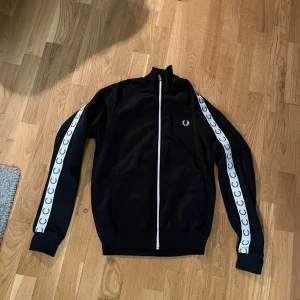Svart track jacket från Fred Perry - Snygg svart track jacket från Fred Perry med vita detaljer och logotyp längs ärmarna. Jackan har dragkedja framtill och är långärmad. Perfekt för en sportig look eller avslappnad stil.