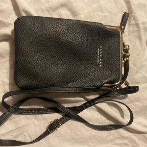 Svart axelväska från Carhartt - Snygg svart axelväska från Carhartt i skinn med justerbar axelrem. Väskan har en praktisk metallram och flera fack inuti för enkel organisering. Perfekt för att bära dina nödvändigheter med stil.