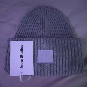 Acne Mössa - Snygg grå ribbad mössa från Acne Studios med deras ikoniska ansiktslogga framtill. Perfekt för att hålla dig varm och stilren under kyliga dagar.