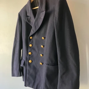 M/1872 Vapenrock – Marinens klassiska "Filtjacka"!  - M/1872 Vapenrock – Marinens klassiska "Filtjacka" (utan gradbeteckningar)   Nu har du chansen att äga en bit av svensk militärhistoria!   M/1872 vapenrock, även känd som den ikoniska "filtjackan", är en tjock, mörkblå filttygsjacka som bars av marinofficerare och specialister som läkare och reservofficerare under 80-talet.   Denna unika jacka, med sin karakteristiska design är ett måste för samlare, militärintresserade eller den som söker ett stilfullt 80-tals vintageplagg med en histor