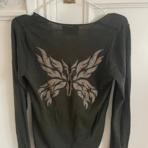 Olivgrön tröja med fjärilsdetalj - Snygg olivgrön tröja från Zadig med en iögonfallande fjärilsdetalj i strass på ryggen. Tröjan har en v-ringad hals och långa ärmar, perfekt för en stilren look.