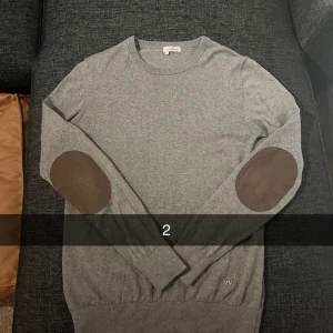 2 Grå tröja från J.Lindeberg - Säljer en stilren grå tröja från J.Lindeberg i storlek M. Tröjan har bruna armbågslappar och ett diskret märke på framsidan. Tillverkad i en blandning av bomull och ull för extra komfort.