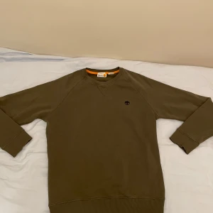 Grön tröja från Timberland - Säljer en grön tröja från Timberland med rund halsringning och broderad logotyp på bröstet. Tröjan har långa ärmar och ribbade muddar vid ärmslut och nederkant. Perfekt för en avslappnad stil. Tröjan är i bra skick.