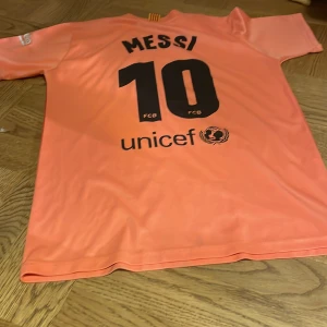 FC Barcelona fotbollströja med Messi 10 - Säljer en cool FC Barcelona fotbollströja i orange och rött med Messi 10 på ryggen. Tröjan har korta ärmar och klubbens emblem på bröstet. Perfekt för fans av Messi och Barca! 🧡❤️