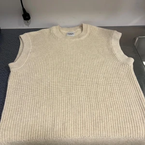 Beige stickad dobber  - Säljer en stilren beige stickad väst med ribbad struktur. Perfekt för lager-på-lager under kyligare dagar. Västen har en klassisk rund halsringning och är ärmlös, vilket gör den lätt att matcha med olika outfits. Västen/tröjan har en liten defekt vid nedre delen vilket gör att priset är nedsatt