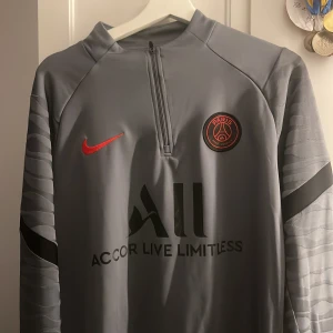 Grå träningsjacka från Nike - Snygg grå träningsjacka från Nike med PSG-logga och röd Nike-swoosh på framsidan. Jackan har en halv dragkedja och texten 'Accor Live Limitless' samt 'Visit Rwanda' på baksidan. Perfekt för fotbollsträning eller som en sportig vardagslook.