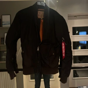 Svart bomberjacka från Alpha Industries - Säljer en klassisk svart bomberjacka från Alpha Industries med orange foder och den ikoniska 'Remove Before Flight'-taggen. Jackan har dragkedja och ribbade muddar vid ärmslut och nederkant. Perfekt för en cool och avslappnad stil.