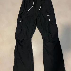 Svarta byxor från Dope - Snygga svarta byxor från Dope med praktiska fickor och snörning i midjan. Tillverkade i 100% polyester, perfekta för utomhusaktiviteter. Byxorna har en stilren design med knappar och dragkedja. Års modell 2022 nypris: 1599 mycket fint skick