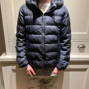 Mörkblå pufferjacka från GANT - Säljer en snygg mörkblå pufferjacka från GANT. Jackan har en dragkedja framtill och en huva för extra värme. Perfekt för kyligare dagar. Skick är 9/10 alltså knappt använda. Skriv om du har frågor eller funderingar. Storleken är 176 men passar Xs-S