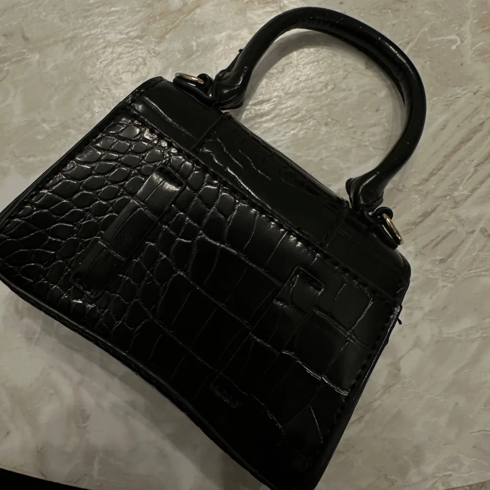 Snygg svart handväska i skinn med krokodilmönster och ett stort guldfärgat B-spänne framtill. Väskan har ett handtag och stängs med en tryckknapp. Perfekt för att ge en elegant touch till din outfit.. Laukut & Käsilaukut.