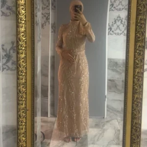 Beige glittrig långklänning - Elegant beige långklänning med glittriga detaljer och långa ärmar. Klänningen har en hög hals och ett bälte i midjan för en smickrande passform. Perfekt för festliga tillfällen.