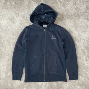 Woolrich hoodie - Säljer en snygg hoodie från Woolrich med dragkedja och broderad logga på bröstet. Tröjan har långa ärmar och en praktisk huva. Perfekt för kyliga dagar!