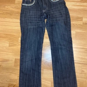 Mörkblå jeans med broderade detaljer - Snygga mörkblå jeans med unika broderade detaljer på fickorna. De har en klassisk straight passform och är perfekta för en avslappnad stil. Jeansens design ger en cool och trendig look. 