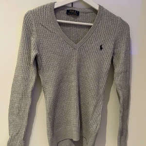 Grå ribbad tröja från Ralph Lauren - Säljer en stilren grå ribbad tröja från Ralph Lauren med v-ringning och det klassiska logotypbroderiet på bröstet. Perfekt för en elegant och avslappnad look.