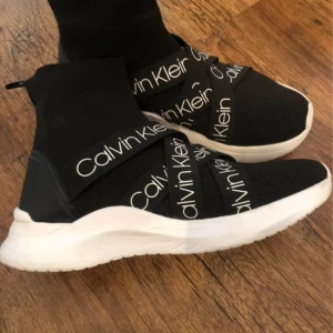 Svarta sneakers från Calvin Klein - Snygga svarta sneakers från Calvin Klein med vit sula och elastiska band med logotyp. Skorna har en modern design med hög ankel och är perfekta för en stilren look.
