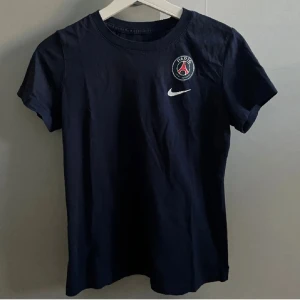 Svart t-shirt från Nike med PSG-logga - Snygg  mörkblå t-shirt från Nike med Paris Saint-Germain-logga på bröstet. Perfekt för fotbollsfans! Kortärmad och i mjukt bomullsmaterial. Använd 1 gång. 
