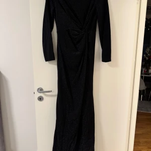 Svart glittrig långklänning - Elegant svart långklänning med glittrande detaljer och en v-ringad omlottdesign. Klänningen har långa ärmar och en stilfull drapering framtill som ger en smickrande passform. Perfekt för festliga tillfällen.