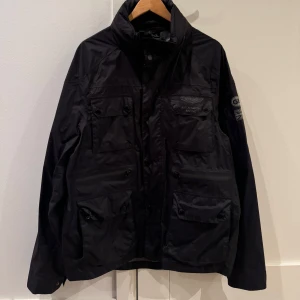 Svart jacka från Hackett - Snygg svart jacka från Hackett med Aston Martin Racing-logga. Jackan har flera fickor och coola detaljer som GB-märke på ärmen. Perfekt för dig som gillar stil och racing.