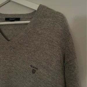 Grå tröja från GANT - Säljer en stilren grå tröja från GANT med v-ringning och broderad logga på bröstet. Tröjan har långa ärmar och ribbade muddar, perfekt för en avslappnad look. Storleken på tröjan säger 2XL, men jag bär storlek M, och tröjan är lite oversized för mig.