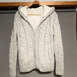 Stickad cardigan jacka med päls - Mysig grå stickad jacka med dragkedja och fodrad insida. Tröjan har ett flätmönster och en stor huva för extra värme. Perfekt för kyliga dagar.