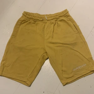 Gula shorts från Quiksilver - Säljer ett par gula shorts från Quiksilver med elastisk midja och en bakficka. Perfekta för en avslappnad stil.