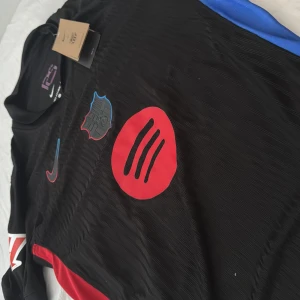 Svart fotbollströja med röda detaljer - Snygg svart fotbollströja från Nike med röda detaljer och klubbmärke på bröstet. Tröjan har korta ärmar och nummer 20 med namnet 'Olmo' på ryggen. UNHCR-logga finns också på baksidan. Den är player version och priset kan diskuteras 