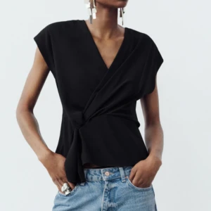 Svart omlottopp - Snygg svart omlottopp med knytning i midjan. Toppen har en elegant v-ringning och är ärmlös, vilket ger en stilren look. Perfekt för att matcha med jeans eller kjol för en chic outfit.