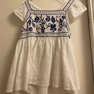 Vit topp med blå broderier från Shein - Charmig vit topp från Shein med vackra blå broderier på bröstet. Toppen har korta ärmar och en knytning i ryggen för en justerbar passform. Perfekt för en somrig look!