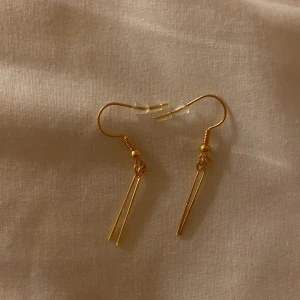 Guldörhängen med minimalistisk design - Snygga guldörhängen med en enkel och elegant design. De har en minimalistisk stil med två smala stavar som hänger från en krok. Perfekta för att ge en stilren touch till din outfit.