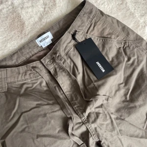 Beige cargobyxor från Weekday - Snygga beige cargobyxor från Weekday med flera praktiska fickor. Byxorna har en avslappnad passform och är perfekta för en casual look. De har dragkedja och knapp framtill. Alldrig använda 