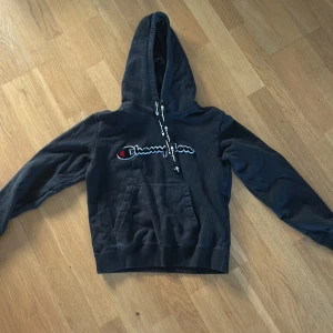 Svart hoodie från Champion - Säljer en svart hoodie från Champion med broderad logga på bröstet och justerbar huva med snören. Perfekt för en avslappnad stil. Den har en praktisk magficka framtill.