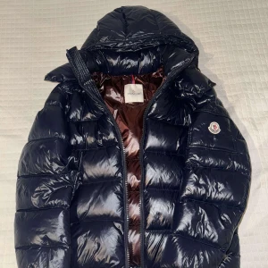 Moncler Maya navy blå  - Unik färg på Moncler jacka med glansig finish och dragkedja. Jackan har en huva och Moncler-logga på bröstet. Perfekt för kyliga dagar. Man kan även scanna märket och det kommer upp moncler officiella hemsida. Finns i storlek S/M samt finns i andra färger som svart