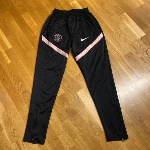 Svarta träningsbyxor från Nike - Snygga svarta träningsbyxor från Nike med rosa detaljer och PSG-logga. Byxorna har en elastisk midja och dragkedjor vid bensluten för extra komfort. Perfekta för fotbollsträning eller annan sportaktivitet.