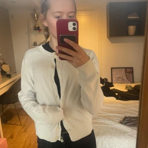 Vit kofta med knappar - Säljer en stilren vit kofta med knappar framtill. Perfekt för lager-på-lager och passar till många olika outfits. Koftan har långa ärmar och en klassisk rund halsringning.