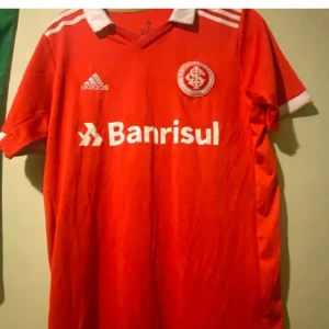 Röd fotbollströja från Adidas - Säljer en röd kortärmad fotbollströja från Adidas med vit krage och vita detaljer. Tröjan har sponsorloggan 'Banrisul' tryckt på både fram- och baksidan samt klubbemblem på bröstet. Perfekt för fotbollsfans!