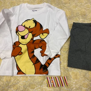 Barn Disney Kläder I 2 Delar  - Säljer en söt vit långärmad tröja med byxor från BabyCoop med ett stort tryck av Tiger från Nalle Puh. Perfekt för barn som älskar Disney-karaktärer. Tillverkad i mjukt100% bomull material för bekväm passform. 5,6 och 7 år kvar.  145kr / 2 delar/ paket 